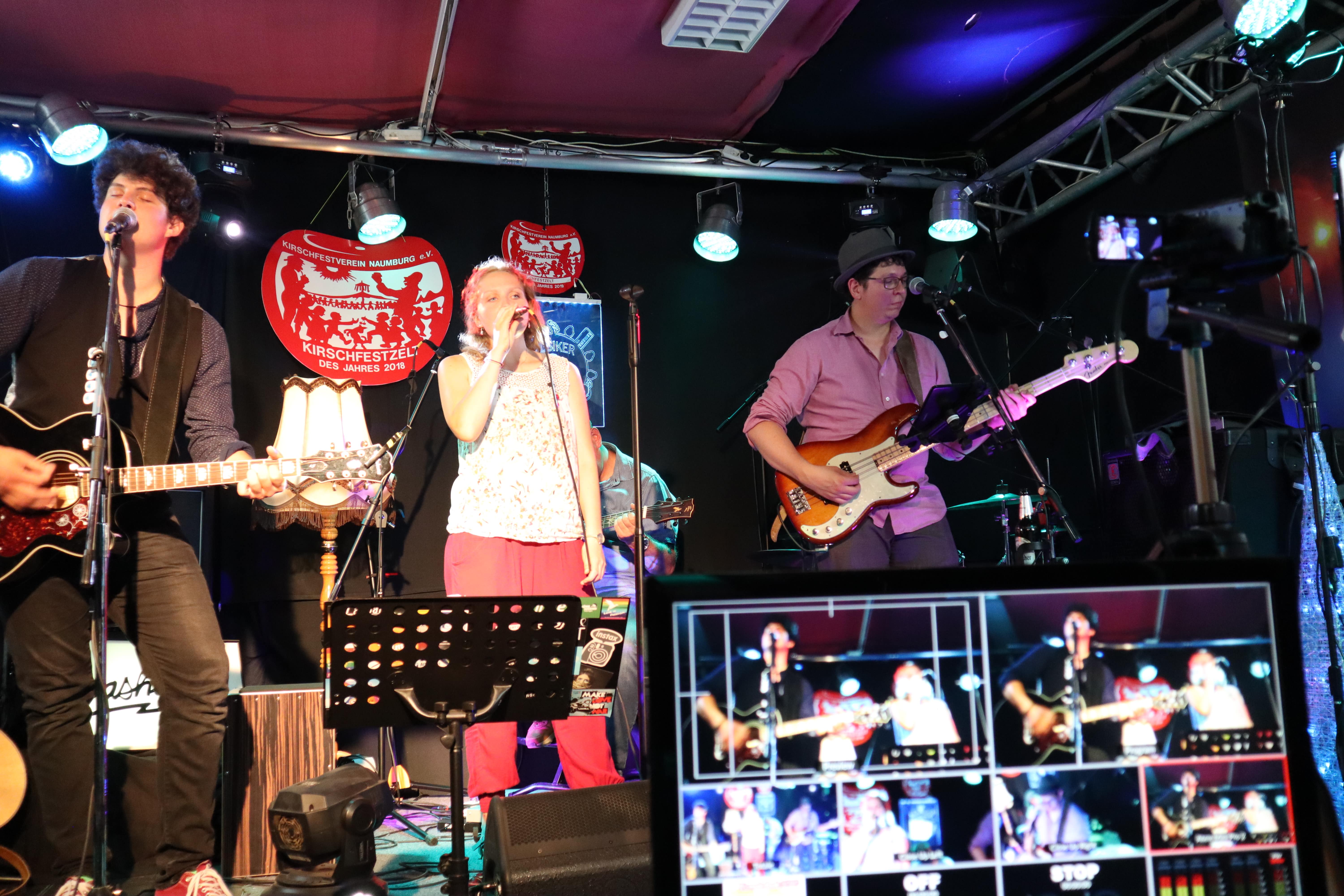 Auf dem Bild ist die Band bei dem Auftritt zum Kirschfest 2020 zu sehen. Tobi singt und spielt Gitarre. Christoph spielt Bass. Charlotte verdeckt Henrik und singt. Im Vordergrund kann man einen Monitor mit verschiedenen Ansichten der Band erkennen. 